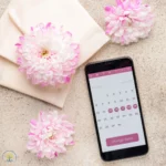 Calendario del ciclo mestruale su uno smartphone, con giorni evidenziati in rosa, posato su uno sfondo rosa con fiori e fogli decorativi.